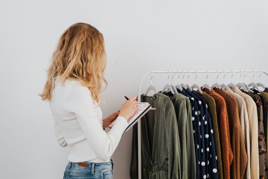Woman writing notes checking a clothing rack with diverse fashion garments indoors.