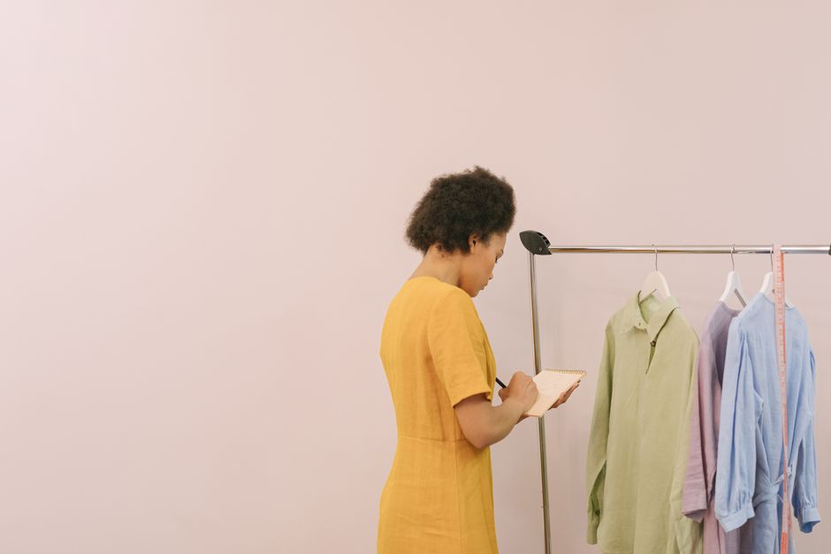 A fashion designer in a mustard dress taking notes beside a clothing rack indoors.